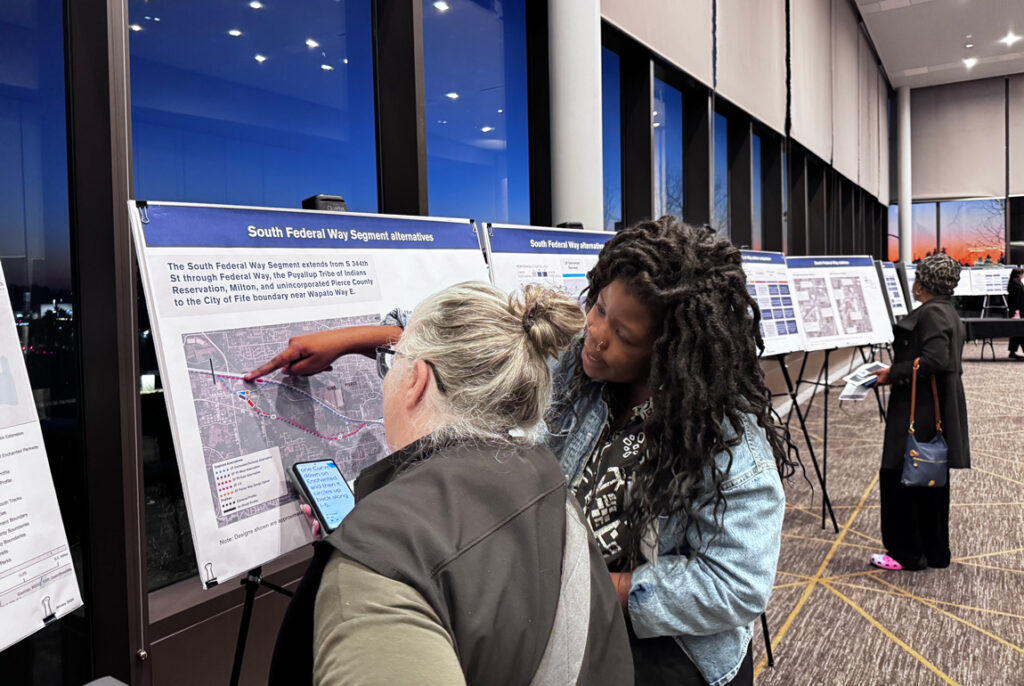 Two people standing in front of a display board map, with one person pointing to a line on the map.